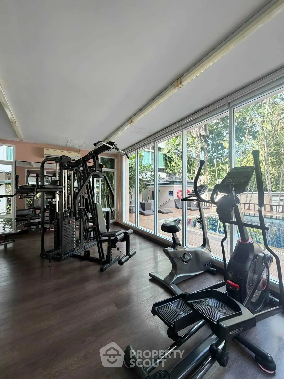 Modern gym with large windows overlooking lush greenery, featuring state-of-the-art exercise equipment.