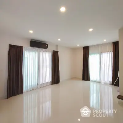 Spacious and well-lit living room with glossy white tiles, large windows draped with elegant brown curtains, and a modern air conditioning unit.