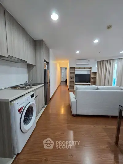 Modern open-plan living room with kitchen and washing machine, featuring sleek wooden flooring and neutral decor.