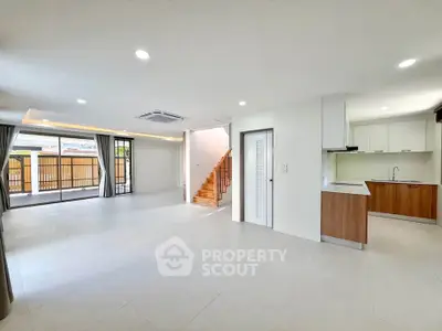 Spacious open-plan living room with modern kitchen and staircase, featuring large windows and ample natural light.
