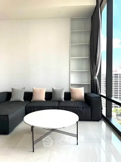 Modern living room with sleek black sofa and large windows offering city views.