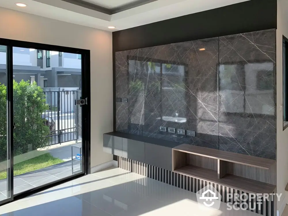 Modern living room with large windows offering ample natural light and a view of the green outdoors, featuring sleek built-in shelving and elegant dark marble wall.