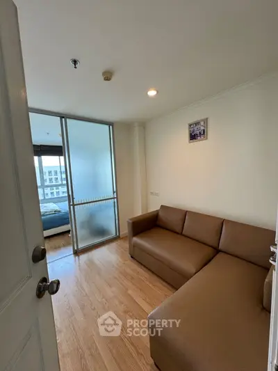 Cozy small living room with brown sofa and wooden flooring, adjacent to a bedroom with glass sliding doors.
