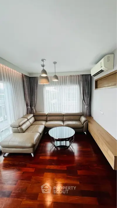 Modern living room with leather sectional sofa, wooden flooring, and large windows with sheer curtains.