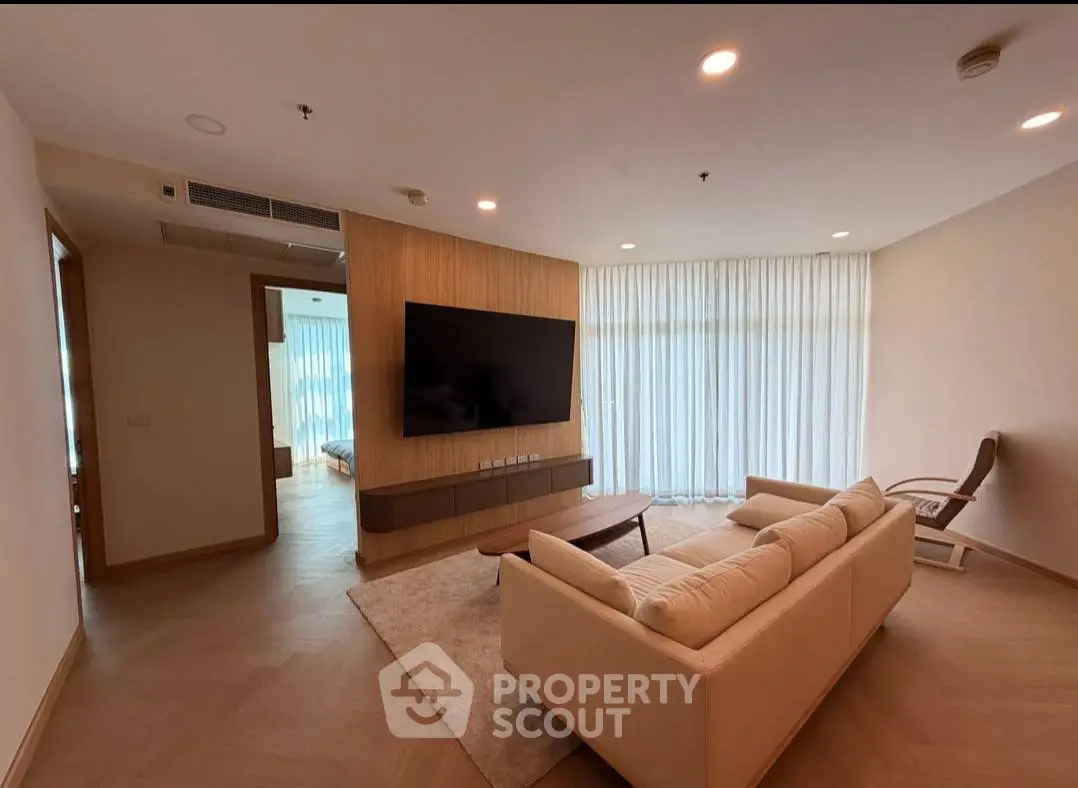 Spacious modern living room with large TV and elegant furniture, featuring natural light from floor-to-ceiling windows.