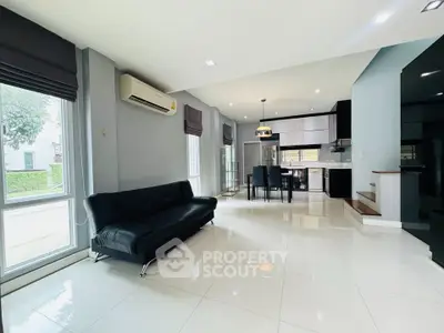 Spacious modern living room with sleek black sofa and open kitchen layout, featuring large windows and glossy tile flooring.