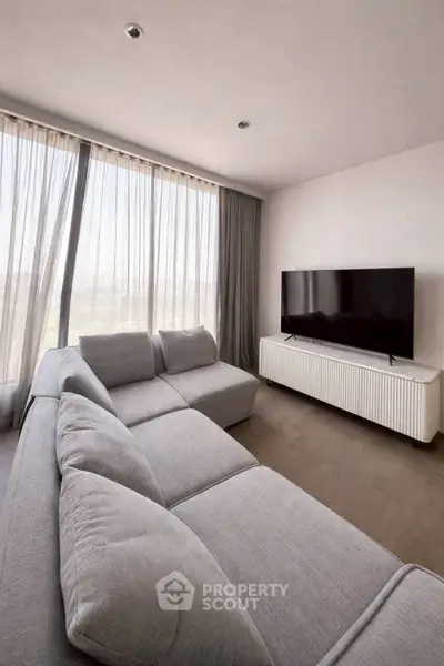 Modern living room with large gray sectional sofa and flat-screen TV, featuring floor-to-ceiling windows.