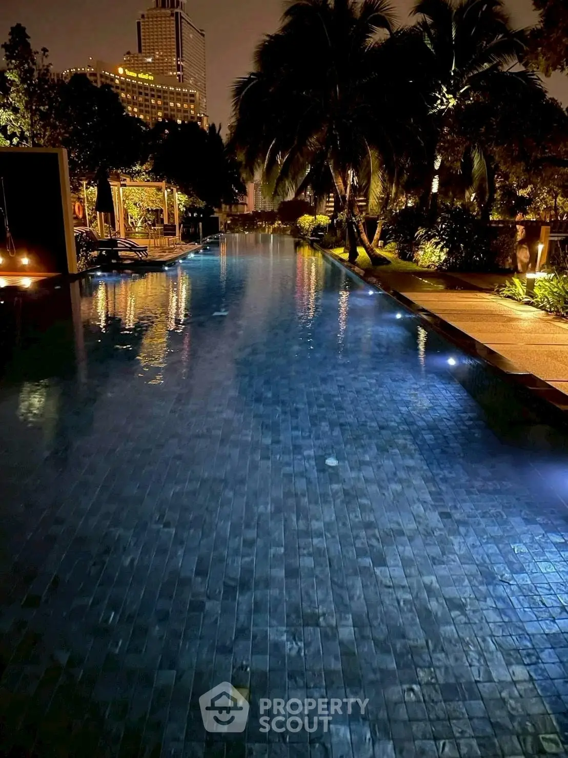 Luxurious night view of a serene swimming pool surrounded by lush greenery and city lights.