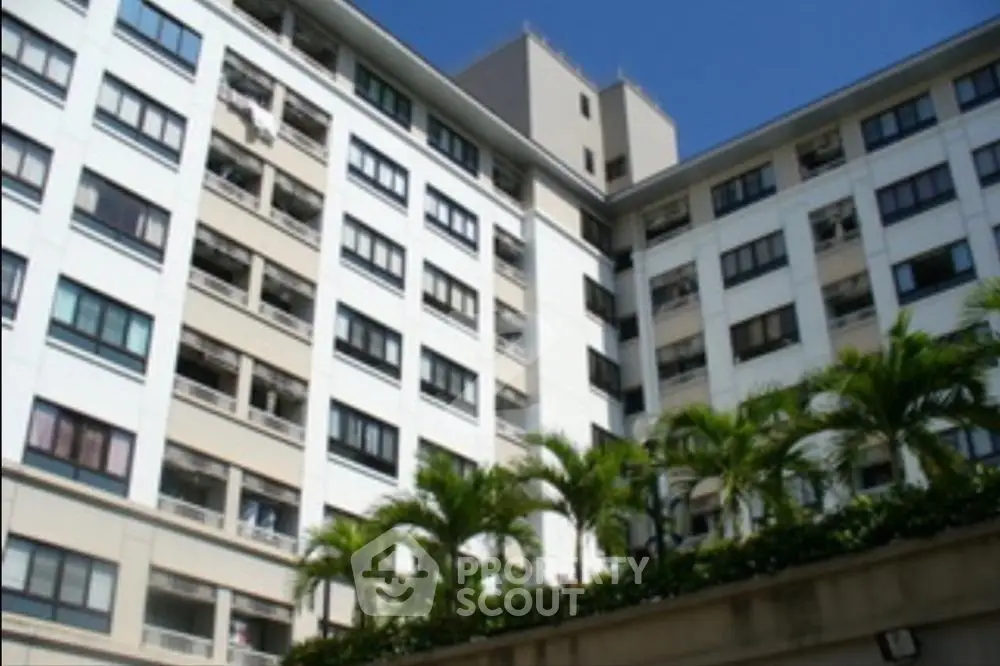 Sunlit modern apartment building with lush greenery, showcasing spacious balconies and a serene urban living environment.