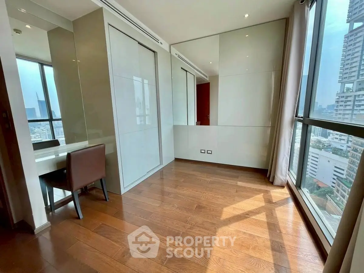 Spacious modern room with wooden flooring and large windows offering city views.