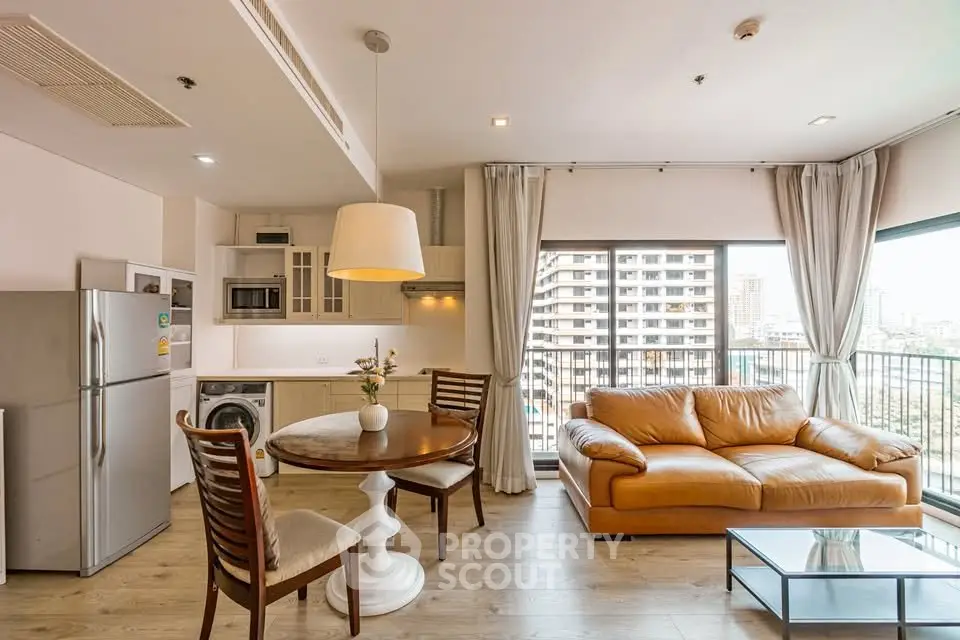 Modern living room with open kitchen, featuring leather sofa and city view from large windows.