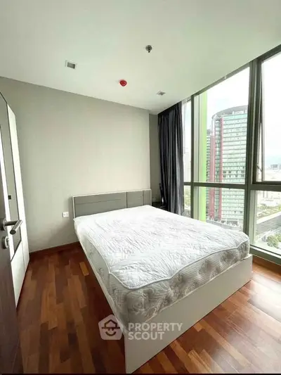Modern bedroom with large windows and city view, featuring a comfortable bed and wooden flooring.