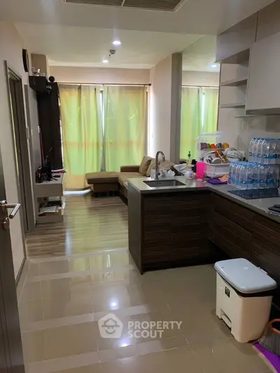 Spacious kitchen with modern cabinetry and glossy tiled flooring opens to a cozy living area, illuminated by natural light from large window with sheer curtains.