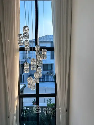 Elegant modern chandelier in a high-ceilinged living room with large windows and a view of the neighborhood.