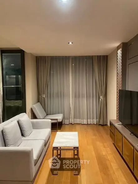 Modern living room with wooden flooring, cozy seating, and large window draped with elegant curtains.