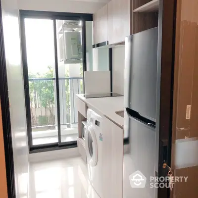 Modern kitchen with washing machine and fridge, bright natural light from large window.