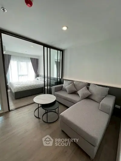 Modern living room with stylish gray sofa and glass partition leading to a cozy bedroom.