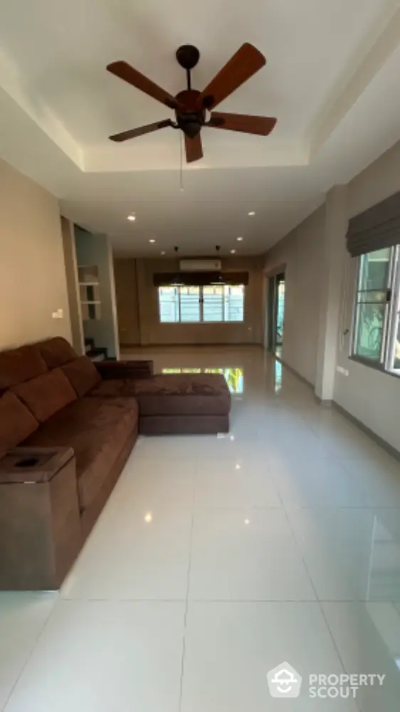 Spacious living room with gleaming white tiles, cozy brown sectional sofa, and a classic ceiling fan, bathed in natural light from large windows.