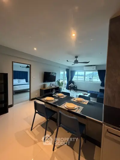 Modern open-plan living room with dining area and bedroom view, featuring sleek furnishings and large windows.