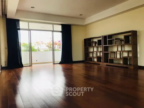 Spacious living room with wooden floors and modern shelving, leading to a bright balcony.