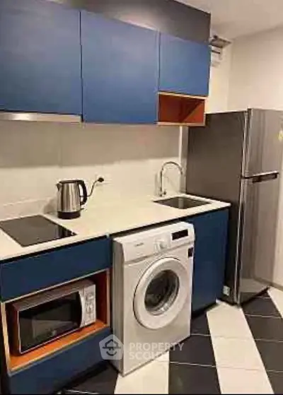 Modern kitchen with blue cabinets, washing machine, fridge, and microwave in compact layout.
