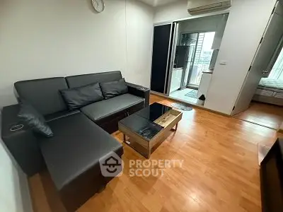 Modern living room with sleek black sofa and glass coffee table, featuring wooden flooring and a glimpse into a bedroom.