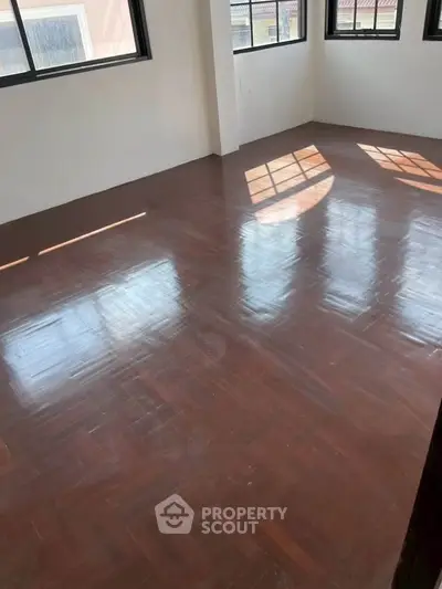 Spacious empty room with polished wooden floors and large windows offering natural light.
