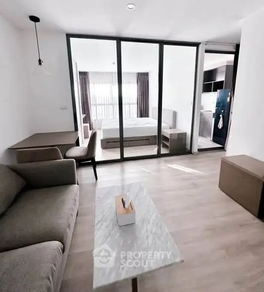 Modern open-plan living room with glass partition to bedroom, featuring sleek furniture and natural light.