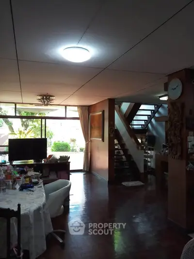 Spacious living room with wooden flooring and staircase, featuring large windows and natural light.
