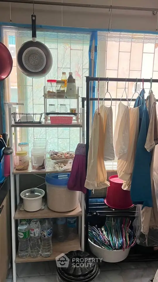 Compact kitchen with hanging clothes, storage shelves, and kitchenware near window.