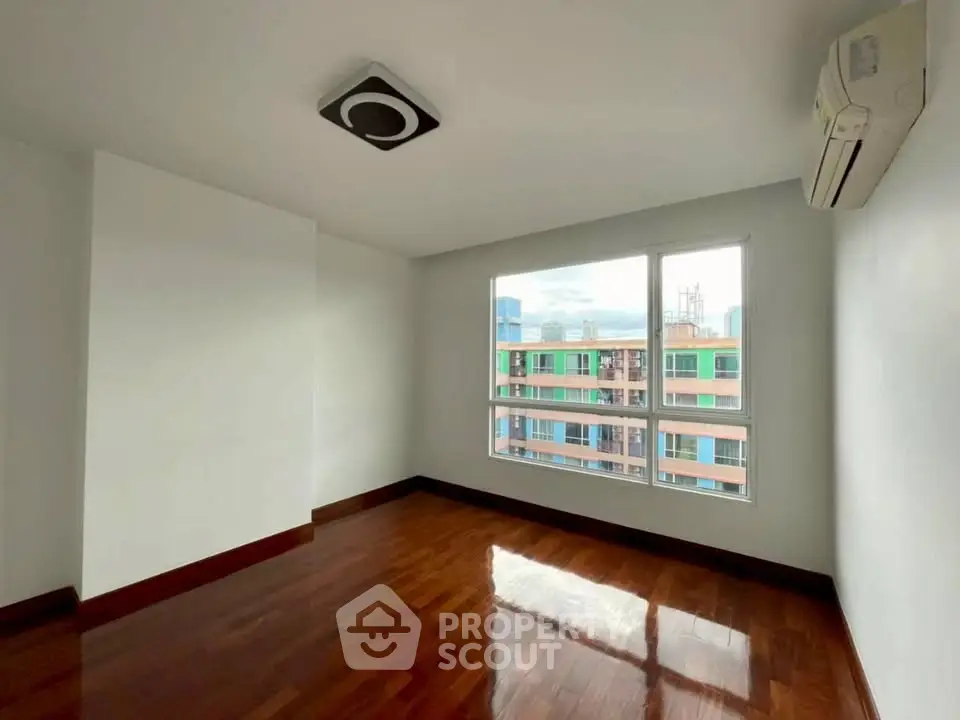 Spacious empty room with wooden flooring and large window offering city view.