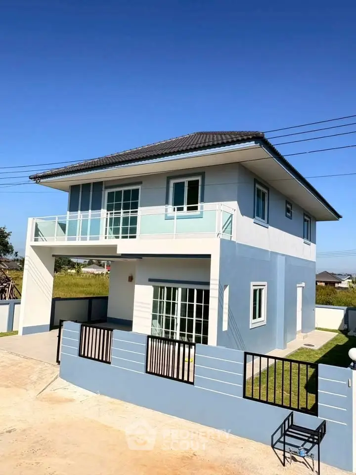 Modern two-story blue house with balcony and gated entrance in sunny neighborhood.