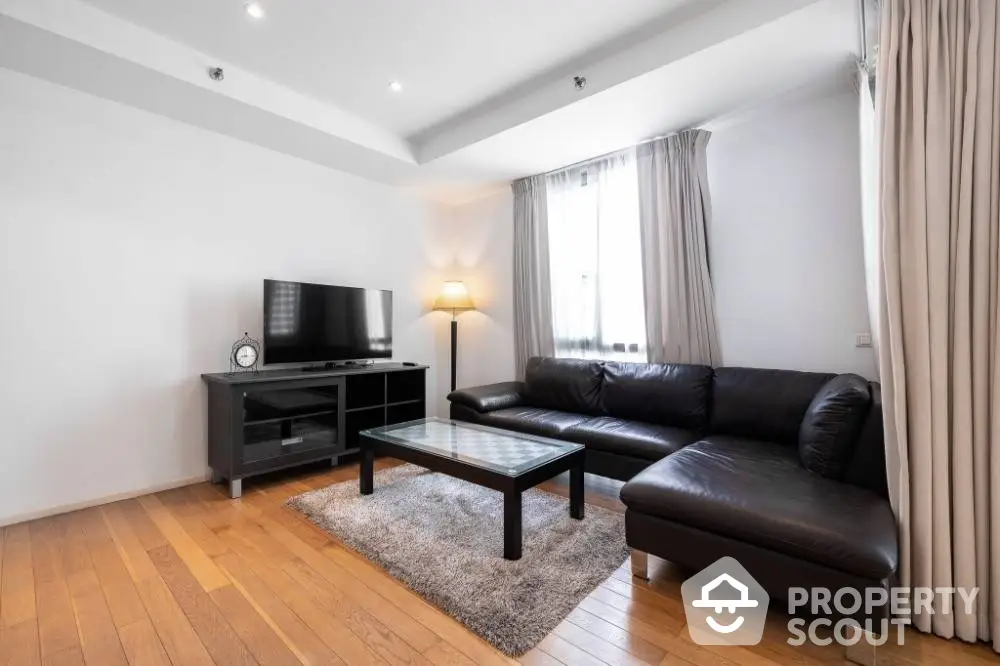 Spacious living room with polished hardwood floors, modern leather sofa, and ample natural light from large window, perfect for relaxation and entertainment.