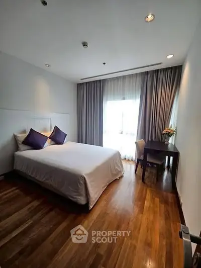 Spacious bedroom with wooden flooring and elegant decor, featuring a large window with natural light.