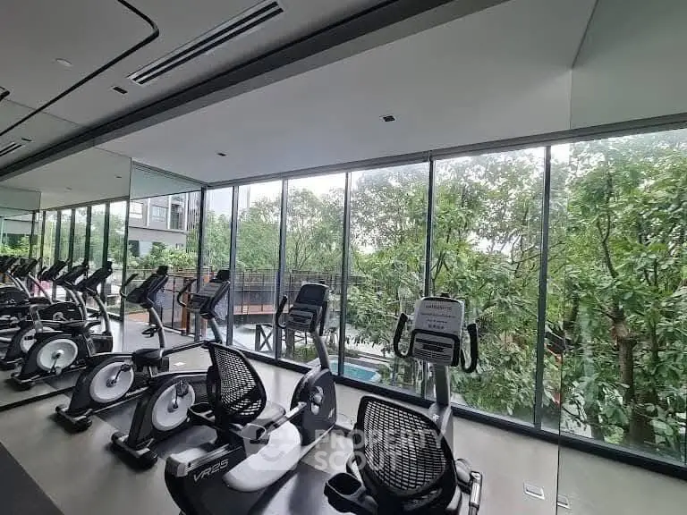 Modern gym with large windows and lush greenery view, featuring state-of-the-art exercise equipment.