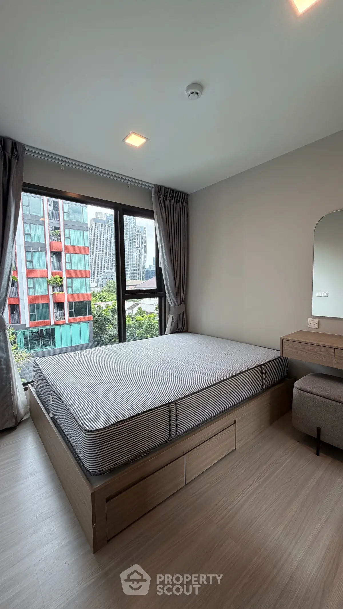 Modern bedroom with large window offering city view, featuring sleek furniture and neutral tones.