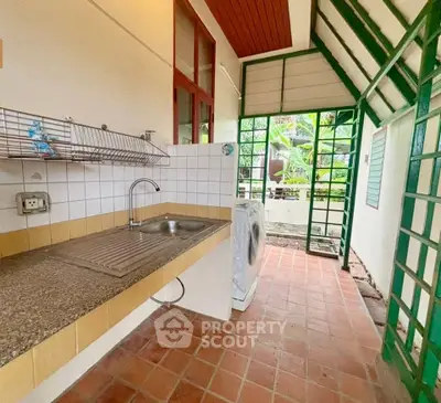 Charming semi-outdoor kitchen with tiled floor and lush garden view, featuring a washing machine and spacious sink.