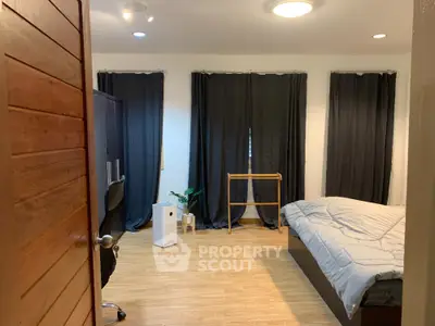 Spacious bedroom with sleek hardwood flooring, modern furnishings, and ample natural light filtering through large windows with dark curtains.