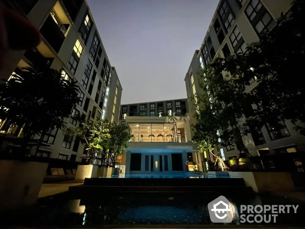 Stunning night view of modern apartment complex with illuminated pool and lush greenery.