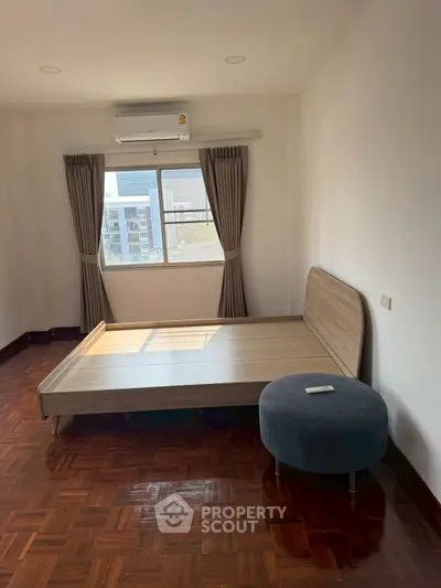 Spacious bedroom with wooden flooring and large window offering city views, featuring a modern bed and air conditioning.