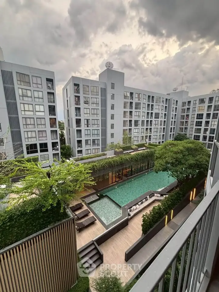 Modern apartment complex with pool and lush greenery under a dramatic cloudy sky.
