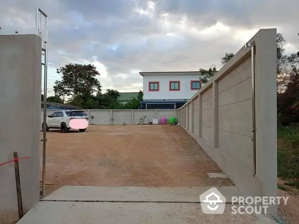 Spacious private parking area within a secure residential compound, featuring a large open gate and a modern two-story house in the background under a serene sky.