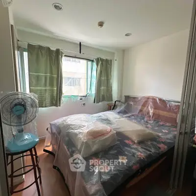 Cozy bedroom with large window and floral bedding, featuring a standing fan and natural light.