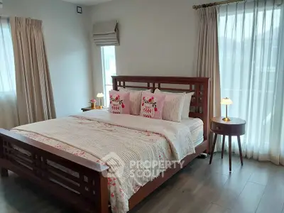 Cozy bedroom with wooden bed frame and elegant decor, featuring soft lighting and large windows with sheer curtains.