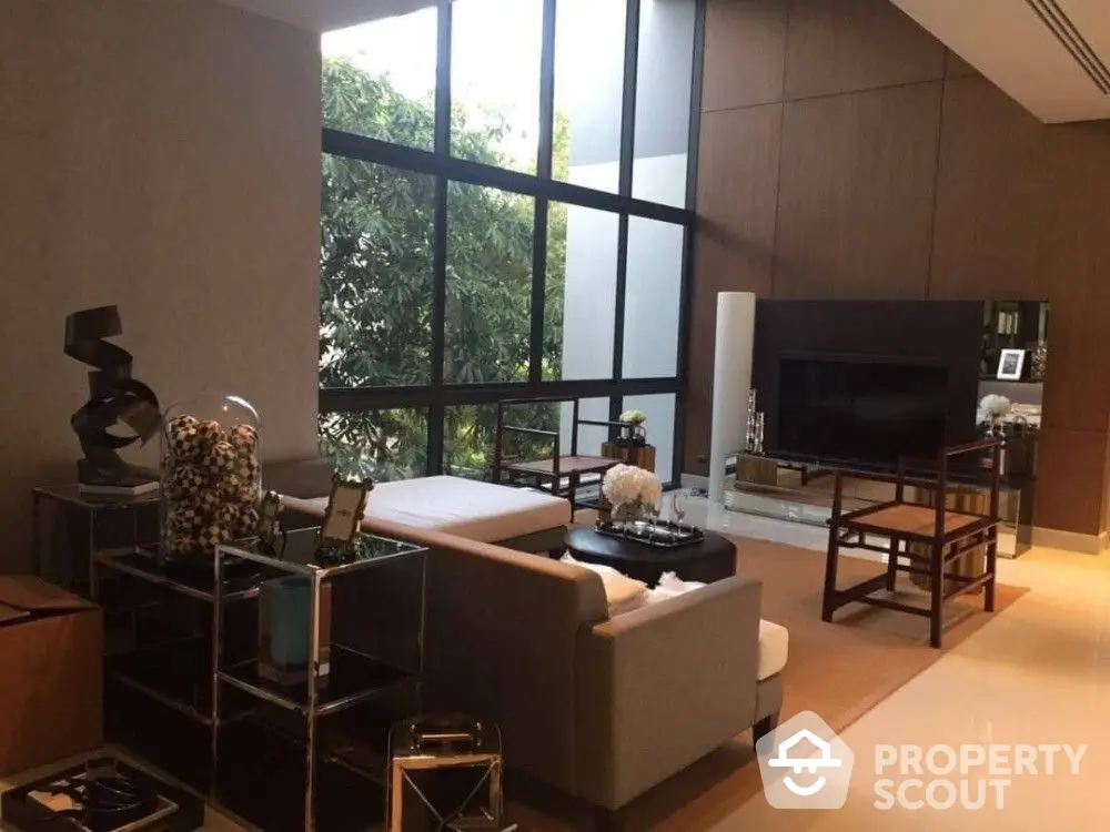 Spacious living room with high ceiling and large windows offering abundant natural light, modern furniture, and a serene view of greenery.