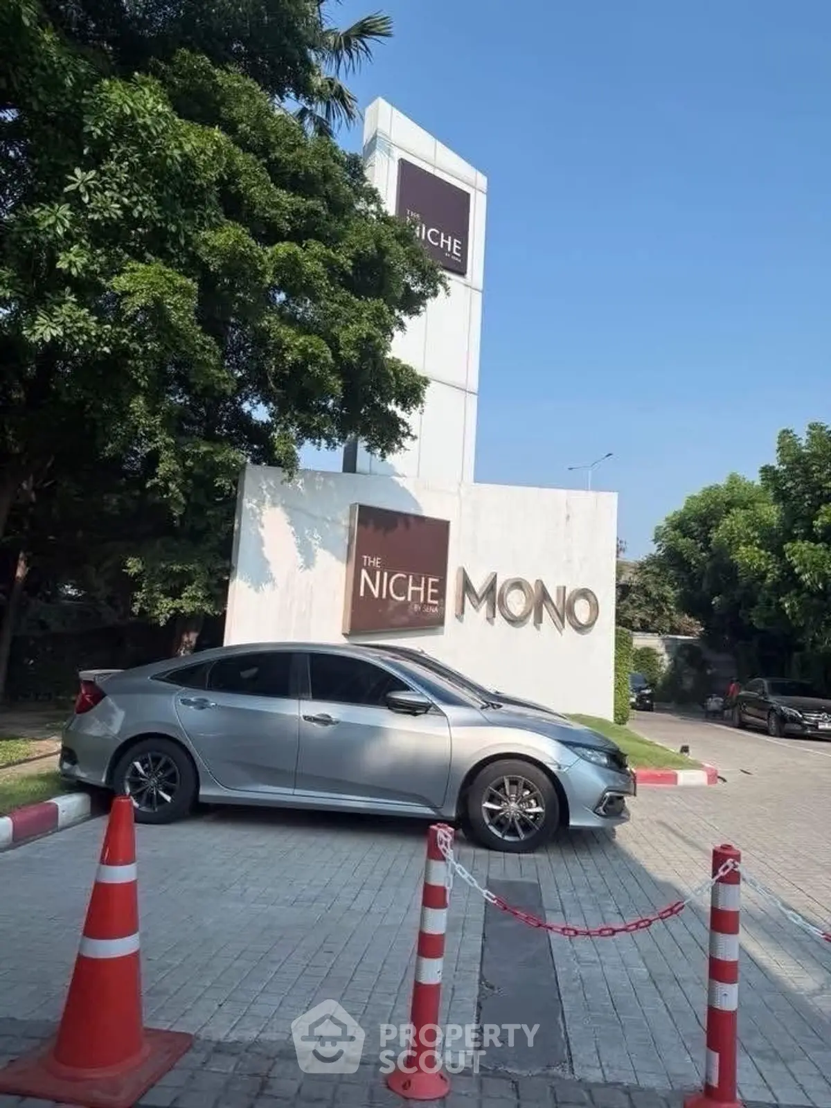 Modern condominium entrance with sleek signage and parked car, surrounded by lush greenery.