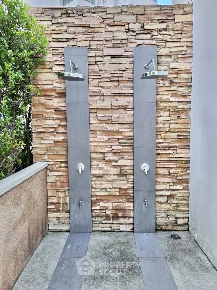 Modern outdoor shower area with dual rain showerheads and stone wall backdrop.