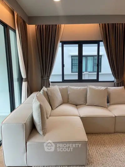 Spacious living room with modern beige sectional sofa and large windows with elegant curtains.