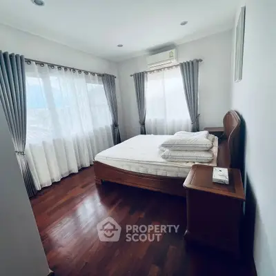 Cozy bedroom with wooden flooring and ample natural light, featuring a comfortable bed and elegant curtains.