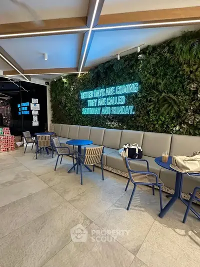 Modern cafe interior with stylish seating and neon sign on a lush green wall, creating a cozy atmosphere.